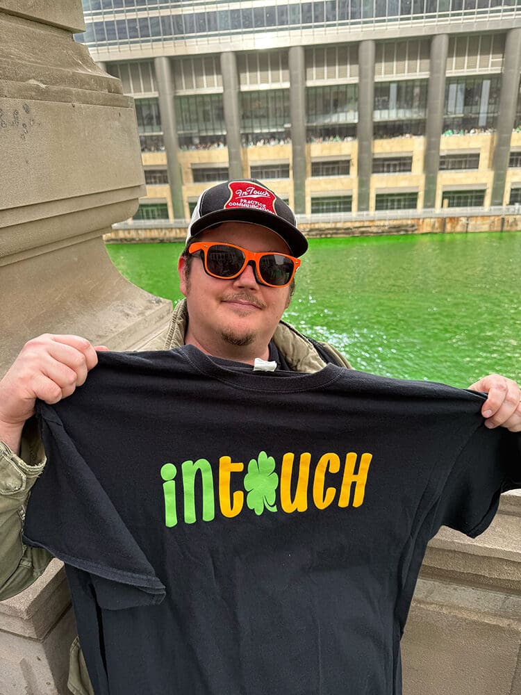 man holding up black intouch st. patrick's day shirt and standing by green river
