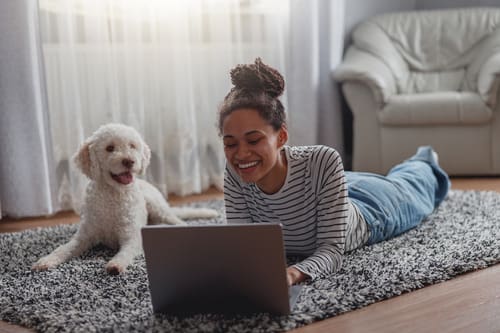 mixed race woman laying on the floor while working on her laptop and smiling with her dog laying next to her